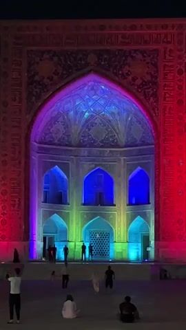 The video shows a night view of the Registan Square in Samarkand, Uzbekistan. The grand archway of one of the madrasahs is illuminated with vibrant, shifting colored lights, primarily in shades of purple, pink, and blue. The intricate tilework and Arabic calligraphy adorning the facade are highlighted by the light display.

In the foreground, several people are visible. Some are standing and looking at the illuminated building, with one person taking a photo. Others are seated or walking, suggesting a relaxed evening atmosphere. The ground appears to be paved, and the overall setting is an open square, indicative of a tourist attraction or historical site.

The time of day is clearly night, as evidenced by the darkness of the sky and the artificial lighting. The weather appears to be clear, as there are no visible signs of rain or wind. The emotion conveyed is one of awe and wonder, with the dramatic lighting transforming the ancient architecture into a spectacular visual display. The location cues, such as the distinct architectural style and the mention of Registan Square, Samarkand, Uzbekistan, pinpoint the specific setting. No prominent text is visible within the video itself.