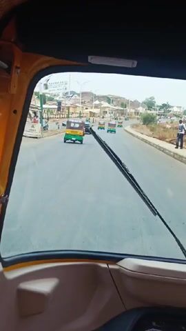 The camera captures a first-person perspective from inside a yellow auto rickshaw, moving along a wide, paved road. The road is light gray, with distinct lanes. The windshield is mostly clear, with a single, dark windshield wiper extending vertically.  Beyond the windshield, the view is of a relatively flat urban or suburban area. Buildings of varying heights and styles line the street.  There are other vehicles on the road, primarily auto rickshaws, similar to the one from which the video is being recorded. There is also at least one sedan-style car.  On the side of the road, there are sidewalks and some trees, but the area appears somewhat arid. The sky is overcast, suggesting a muted, diffused light. There are a few people visible on the sidewalks. The interior of the auto rickshaw is visible in the frame, with yellow and black elements.