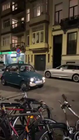 The video shows a teal Fiat 500 driving down a street at night. The car is turning at a crosswalk in a city street. There are other cars, bikes, and businesses on the street. There are people at a restaurant with covered outdoor seating. The buildings are old with many windows.