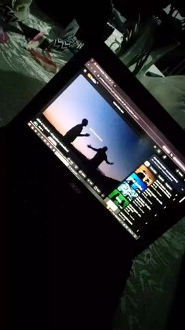 A laptop is on a bed, playing a YouTube video. The video appears to be a music video, with a dark silhouette of two people against a sunset background. The laptop is open, with the screen displaying the YouTube interface, including suggested videos on the right side. The surrounding environment shows bedding and other items in a dimly lit room. The laptop brand is Acer.