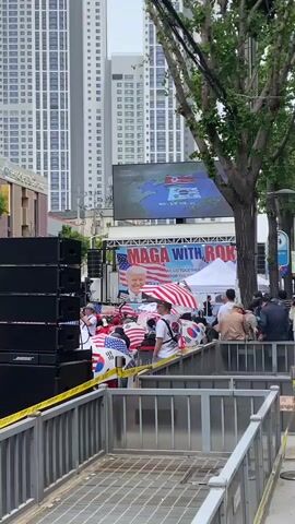 The video shows a political gathering with a "MAGA WITH ROK" banner in an urban setting, possibly in South Korea, given the presence of the South Korean flag. Many attendees are sitting in chairs, holding umbrellas decorated with both American and South Korean flags to protect themselves from the rain. A large screen displays a map with the flags of North and South Korea. Stacks of Bose speakers are positioned on the left side of the shot, suggesting an amplified sound system for speeches or announcements. Yellow police tape marks the perimeter of the event. Tall buildings and trees are visible in the background, establishing a street scene in a city.