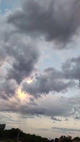 The video shows a cloudy sky at dusk. The clouds are dark and grey, with some lighter patches where the sun is peeking through. The sun is setting, casting a warm, golden light on the clouds. In the foreground, there is a line of trees and some houses. The overall impression is one of a peaceful and serene evening.
