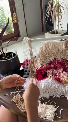 A close-up shot captures a person's hands in the process of creating a tufted wall hanging. The artwork, which appears to be near completion, features a textured design with rows of yarn in cream, beige, deep red, and gold. The tufting is being done on a white mesh canvas stretched over a frame. The person is holding a tufting tool and manipulating cream-colored yarn, working on the lower section of the piece. Loose bundles of yarn in beige and cream are scattered on the wooden table in front of the artist, along with some pre-made fringe.

The setting suggests an indoor environment, possibly a home or studio. Behind the artwork, a window offers a glimpse of lush greenery, indicating a tropical or subtropical climate. A large potted plant with dark leaves is visible to the left. In the background, a tall, slender plant with spiky leaves and reddish-pink hues adds to the vibrant, natural ambiance. The lighting appears to be natural, coming from the window, suggesting it is daytime. The overall mood is one of focused creativity and hands-on craftsmanship. No visible text is present in the media.