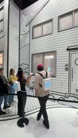 A video captures a small crowd gathered in front of a sizable, cartoonish building facade. The building is rendered in black and white outlines, creating a whimsical, two-dimensional effect. Several people are interacting with a game or display built into one of the windows. Each participant has a small, white handheld device connected by a cord to a stand. They seem to be aiming or interacting with the window display, which shows a glowing blue bar and the word "Rocky?" written above. The people are a mix of genders and races, with one individual wearing a pink hat and another in a full white suit. They seem to be engaged and entertained. The overall environment feels like an exhibition or interactive art installation. The ground has a black and white pattern that enhances the cartoonish aesthetic of the scene.