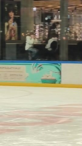 The video shows three people skating on an indoor ice rink. One person in a black jacket and light grey pants is at the left of the frame. A second person in a black jacket and pants skates from left to right across the middle of the frame. A third person in a blue jacket and grey pants skates from left to right behind the second person. Behind the skaters is the rink’s barrier with an advertisement reading “super akropolelatvija.lv, Akropole Alfa, Akropole Riga, dāvana”. Beyond the barrier is the interior of a shopping mall. A poster with a woman’s image is also visible.