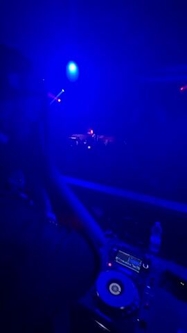 The video shows a DJ playing music at a club. The scene is dimly lit with blue and red lights shining over the crowd of indistinct figures dancing in the background. The DJ stands behind a console with buttons, dials and a CD player.  A water bottle sits to the right of the console. The DJ is moving their hand, presumably to adjust the music playing.
