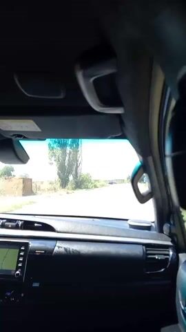The video shows a view from inside a car, looking forward through the windshield. The car is driving on a paved road with vegetation on either side. The dashboard of the car is visible in the foreground, including a navigation screen. In the distance, another car is visible on the road. There are road signs visible ahead. The sky is bright and appears to be daytime. The audio consists of a voice speaking in Bulgarian, discussing road regulations concerning animals and religion.