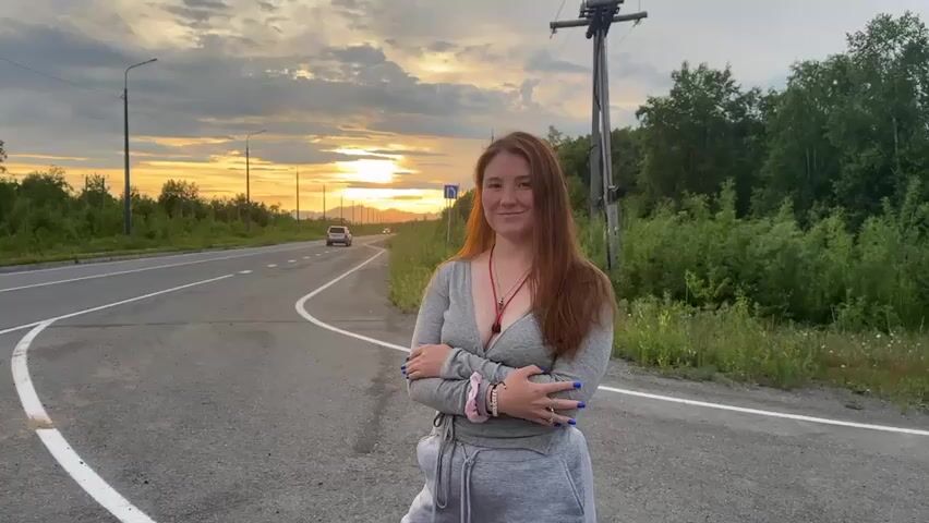 A young woman with long red hair stands at the side of a paved road with a white line marking the edge. She is wearing a grey long-sleeved shirt with a deep V-neck and grey sweatpants. Her arms are crossed in front of her. She has dark blue fingernail polish. There are bracelets on her left wrist. A white bag is hanging from her waist. Behind her, the road curves to the left and continues into the distance where a vehicle is visible. The sky is a mix of clouds and a bright sunset with orange and yellow hues. Trees and vegetation line the side of the road. A utility pole with wires is visible to the right of the woman. The scene appears to be a rural or suburban roadside during sunset. The woman looks directly at the camera and smiles, then turns her head to look out at the sunset over the road.