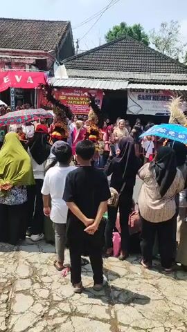 A crowd of people, many wearing headscarves, are gathered outdoors on a paved surface. In the center of the crowd, several individuals are dressed in elaborate costumes, including one person in a tall, feathered headdress. Some people are holding umbrellas. In the background, there are buildings with signs. The sky is visible and appears to be sunny.