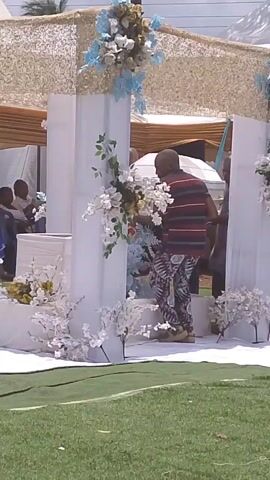 The video shows a decorated outdoor stage, possibly for a wedding or memorial service. The stage is adorned with white and blue floral decorations and has a white fabric covering. People are seen entering and exiting the stage area. They are dressed in traditional and Western clothing. Some people are seated and watching. The environment is sunny with green grass. The sound of music can be heard in the background.