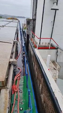 The video shows a ship entering a lock chamber. The ship is moving slowly forward into a narrow channel. The walls of the lock chamber are high and made of concrete. There are ropes and railings visible on the ship. The water level in the lock chamber appears to be lower than the surrounding water. The sky is overcast and the overall lighting is dim. There are some floating structures visible in the distance to the left. The perspective is from the deck of the ship, looking forward and slightly to the right. The camera operator is speaking in Russian, commenting on the ship's movement.