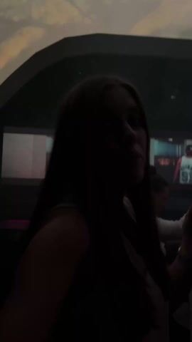 The video, shot in very low light, shows a young woman in a dark room, possibly a nightclub or bar. She is facing the camera, smiling slightly, and appears to be holding a phone or small device in her hand. The room has colored lights (pink and purple) along the walls. Screens are visible in the background, showing moving images. The overall atmosphere is dark and noisy, possibly due to music playing.