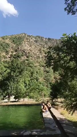 The video shows an outdoor swimming pool with green water surrounded by a concrete edge. Two young men are sitting on the edge of the pool, with their legs hanging over the side. They are looking towards a yellow slide that is set up next to the pool. The background of the scene is a lush green hillside covered in trees, with a rocky mountain peak visible at the top. The sky is blue with a few white clouds. The overall atmosphere is peaceful and recreational.