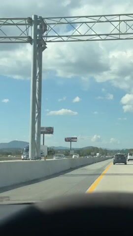This video shows a view from the driver's seat of a vehicle traveling on a highway. The sky is blue with white clouds. There are two billboards visible on the left side of the highway. The first billboard has an advertisement for "FULL PLATES." The second billboard has an advertisement that is mostly obscured by clouds but appears to be for a car insurance company. Traffic is visible on the highway, including cars, trucks, and vans. The vehicle is moving at a steady pace. The time stamp indicates the video is about 4 seconds long.