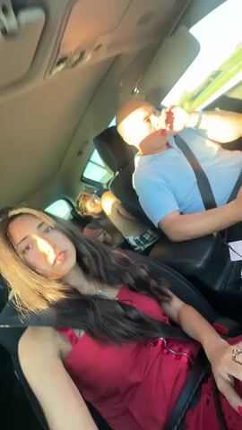 The video shows four people in a car. A woman in a red dress is in the front passenger seat, looking at the camera. A man with a shaved head and a blue shirt is in the driver's seat, looking forward. Two young men are in the back seat. One is asleep, and the other is looking forward. The car appears to be moving. The sun is shining.