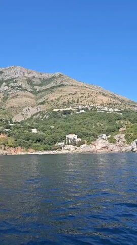 A boat is moving through blue water toward a coastline with a steep, rocky mountain in the background. The mountain is covered in patches of green vegetation and dotted with white buildings. The coastline has cliffs and a small beach area with people and lounge chairs. The sky is clear and blue.