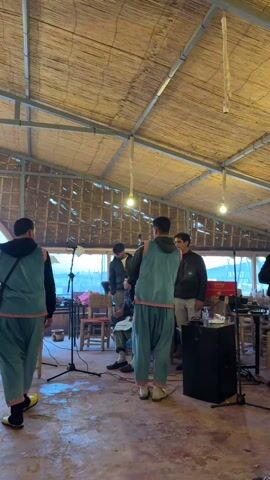 Inside a rustic, high-ceilinged structure with a bamboo roof, a group of people are gathered, some seated and others standing. Two men dressed in matching teal tracksuits with neon orange trim stand center stage, facing the group. In the foreground, a hand in a white sweater holds a decorated hat with tassels. The view shifts to capture the audience raising their hands in the air, creating a sense of participation. A small fire burns in a metal container. The walls of the structure are partially open, revealing a view of the outside.