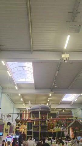 The video captures the interior of a bustling indoor playground, teeming with people, mainly adults and children. The space is brightly lit by overhead fluorescent lights and natural light filtering through skylights in the gray, corrugated metal ceiling. At the center of the frame, there's a large, multi-level play structure with tunnels, slides, and other play elements, topped with a giant Viking helmet. Numerous people are gathered around tables on the floor level, some appear to be eating and socializing. In the background, additional play equipment is visible. The camera is static, providing a wide view of the crowded indoor play area.