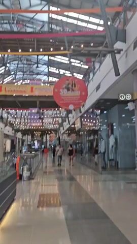 The video shows the interior of a shopping mall with a high, exposed ceiling structure. Banners advertising sales and promotions are visible. People are walking through the mall. A model of three high-rise buildings is displayed in the foreground. The model shows the buildings in detail with illuminated windows and landscaping. The buildings are white. The model sits on a dark grey base with glass surrounding the model.