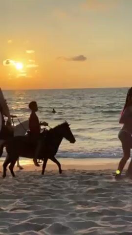 The video depicts two children riding ponies on a beach at sunset. One pony is dark brown and the other is white.  The children wear dark shirts, and the sky is a golden-orange color as the sun sets over the ocean. Waves break on the shore, and a few people swim in the distance. The ponies walk along the sand, led by an adult whose legs are visible. A yellow ball sits on the sand in the foreground.