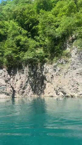 The video shows a person kayaking on a vibrant turquoise lake. The camera is positioned behind the kayaker, showing their hands on a paddle as they move through the water. The lake is surrounded by steep, rocky cliffs covered in lush green trees. Other kayakers can be seen in the distance, also navigating the water. The sky is a clear blue. The scene appears to be a scenic outdoor activity in a natural environment.