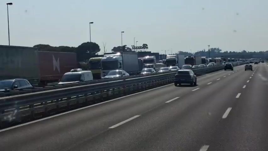 The video shows a busy highway with a significant traffic jam. Numerous cars and trucks are stopped or moving slowly in multiple lanes. On the left side of the highway, there's a median barrier, and beyond that, more vehicles are visible. In the background, there are some buildings and trees, suggesting an urban or suburban setting. The sky is clear and bright, indicating daylight. There are no people clearly visible in the video, and no specific actions or interactions are taking place other than the movement of vehicles. A sign with a warning symbol is visible on the left side of the highway.