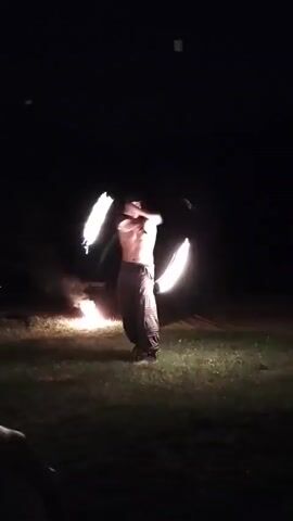 A young male, shirtless and wearing dark pants, is performing with fire sticks in a dark outdoor setting. He is standing in a grassy area with a bonfire to his left. He is holding two flaming sticks, one in each hand, and appears to be juggling them in a circular motion. The fire from the sticks and the bonfire illuminates the scene. There are faint shapes that could be people or objects in the background, but they are not clearly defined due to the darkness. The video is short and captures a brief moment of the performance.