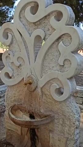 A close-up shot focuses on a decorative stone fountain with water flowing from a brass spigot into a carved basin. The fountain features ornate, swirling carvings above the spigot. The water runs down the rough-textured stone below the basin, disappearing out of frame. The background is blurred, suggesting an outdoor environment with some greenery and possibly trees.