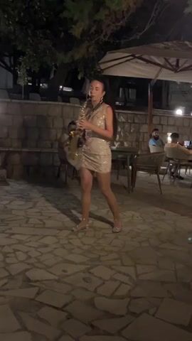 A woman in a sparkly dress plays a saxophone at an outdoor venue at night. She stands in the center of a paved area, with tables and chairs for patrons visible in the background. She is facing the viewer and her hair is tied back in a ponytail. The lighting is dim, with some overhead lights illuminating the scene. A stone wall and trees are visible behind the woman.
