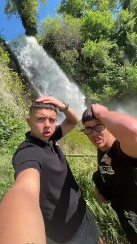 Two young men take a selfie in front of a tall waterfall. The man on the left wears a black polo shirt and black shorts, while the man on the right wears a black t-shirt with a graphic design and glasses. Both have their hands on their heads. The waterfall rushes down a rocky slope, surrounded by lush green foliage under a sunny blue sky.