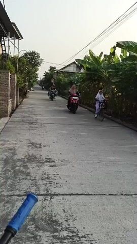 The video shows a first-person perspective of someone on a bicycle or motorcycle. They are riding down a paved road in a residential area. There are several other people on motorcycles and one person on a bicycle ahead of them. The road is lined with houses, trees, and plants. The sky is clear and the sun is shining. The person filming is moving forward, and the people ahead are also moving forward. The people on motorcycles appear to be going about their daily business, some with passengers or cargo. The scene suggests a typical day in a neighborhood.