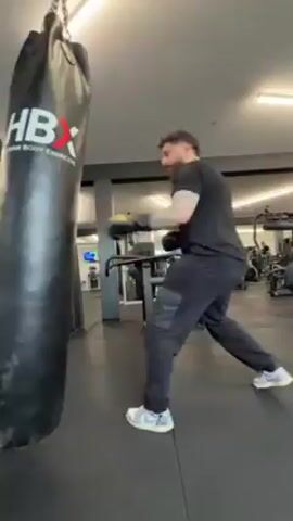 A man wearing black clothing and boxing gloves is punching a black HBX heavy bag hanging from the ceiling. He moves around the bag, alternating between throwing punches and ducking to avoid being hit by the bag's swinging motion. The scene is set in a gym, with various exercise machines visible in the background. The floor is black rubber.  The lighting is bright. The man has short dark hair and a beard. He has a tattoo on his left arm. He wears white sneakers.