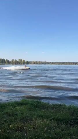 A person is riding a yellow and white jet ski on a body of water. The jet ski is moving from left to right in the frame, creating a wake behind it. The water is calm with small waves. In the background, there is a shoreline with trees. The sky is blue and clear. The foreground shows grass and some fallen leaves. The video is shot from a low angle, near the water's surface.