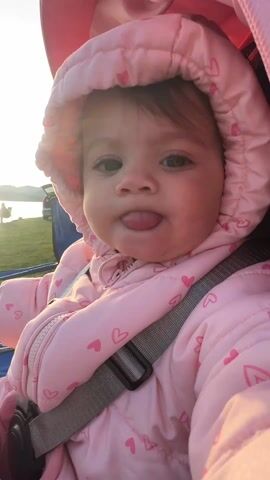 The video shows a close-up of a baby in a pink snowsuit with hearts, strapped into what appears to be a stroller. The baby sticks their tongue out and then stares into the camera with a slight pout. The background shows glimpses of grass and a blue object. The baby has brown hair and eyes. The hood of the snowsuit is up.