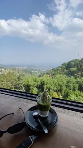 The video displays a close-up shot of a dessert in a glass, placed on a black saucer with a spoon. The dessert appears to be layered, with a green base and a lighter colored, swirled topping, possibly ice cream or a mousse. Sunglasses and a vape pen are visible on the wooden surface in the foreground. In the background, through a large window, a panoramic view of lush green hills and a distant town under a bright blue sky with fluffy white clouds is seen. The lighting suggests it is daytime. There are no people visible in the video, and no actions or interactions are taking place.