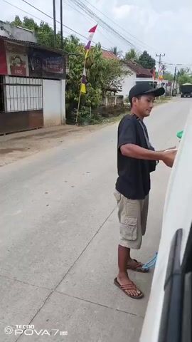 A person wearing a black t-shirt, khaki cargo shorts, a black baseball cap, and brown sandals is standing on the side of a paved road. They are holding a stack of what appear to be bottled drinks in their right arm and hand, and are in the process of placing them into the open back of a white vehicle. A blue rope or strap is visible near the person's feet, attached to the vehicle. In the background, there are buildings, trees, and utility poles. There are also several flags displayed on poles. The scene appears to be outdoors during the daytime, with a cloudy sky.