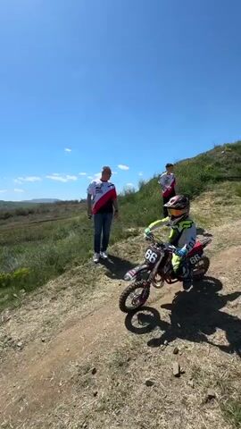 A child in motocross gear is on a small dirt bike, revving the engine. The background shows a grassy area and a clear blue sky, with rolling hills in the distance. The child starts riding the bike, moving along a dirt path. Two adults are standing by the side of the path, watching the child. In the background, a fenced area with vehicles and structures is visible, along with snow-capped mountains in the far distance.