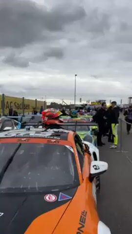 The video shows a line of race cars, some with people standing around, on a cloudy day. The camera pans along the cars, showing different colors and designs. Most are Porsches. Two people in racing suits are talking next to a white and blue car with an Israeli flag on it. A helmet sits on top of the car. In the background, there are signs that say "ADAC Sachsen".