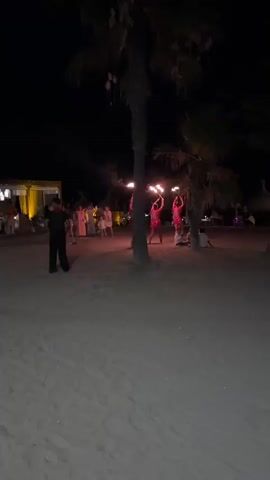 The video captures a lively evening scene on a beach, likely at Playa Del Sol-Villacana, Spain. The setting is a sandy beach at night, illuminated by warm artificial lighting from a nearby building and the flickering glow of fire torches. 

The central focus is a group of performers, possibly dancers, who are skillfully twirling torches ablaze with fire. They are dressed in vibrant, eye-catching outfits, adding to the energetic atmosphere. A crowd of spectators is gathered around, some standing and watching the performance, while others are seated on the sand. Among the audience are children, who appear to be captivated by the spectacle.

The overall mood is festive and celebratory, with the beach setting and the fire dancing creating a magical ambiance. The presence of palm trees further enhances the tropical, holiday feel of the location. The time of day is clearly night, as indicated by the dark sky and the artificial lighting. The weather appears to be clear, suitable for an outdoor event.

In the background, a building with illuminated windows suggests a restaurant or event venue. The sand itself is textured, with footprints visible, indicating recent activity. The scene is dynamic, with the performers in motion and the crowd engaged, creating a sense of lively entertainment.