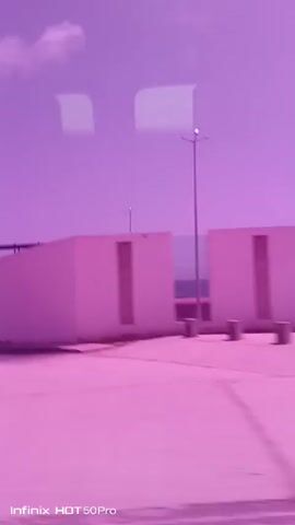 This is a video of passing scenery, likely from a moving vehicle, with a distinct purple-pink color cast. The scene appears to be an urban or industrial area under a bright sky with scattered clouds.

The subjects are primarily buildings and infrastructure. We see a long, low-slung white building with dark rectangular openings, possibly windows or doorways. There are also areas of exposed metal framework, suggesting construction or industrial structures. Some of these structures have a tiled or paved ground in front of them. In one frame, a light pole is visible. In another, a small white car is parked near a building entrance. There are also glimpses of what appear to be trees and shrubbery.

The activity is passive observation of the passing environment. The motion suggests travel.

Notable details include the bright, possibly sunny, time of day due to the clear sky and clouds, though the color cast makes precise determination difficult. The presence of streetlights and what seems to be a police building ("POLICIA" is visible in text) suggest it is a populated area. The overall mood is neutral, with no strong emotional cues conveyed by the imagery.

The visible text reads "Infinix HOT 50 Pro" at the bottom of the frame, indicating the device used to record the video.