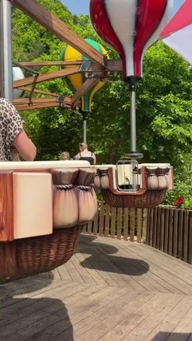 The video shows a children's amusement park ride with several gondolas shaped like hot air balloon baskets. The gondolas are attached to an overhead rotating arm and are moving in a circle. Each gondola has people in it. In one gondola, there is a woman with a tattoo on her back and two young girls; one girl is wearing a blue hat and holding a stuffed animal.  The ride is on a wooden deck surrounded by a low wooden fence, and there are green trees in the background. The weather is sunny.