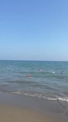 This media file captures a lively scene at a beach on a clear, sunny day, likely in the afternoon given the bright light and blue sky. The setting is a sandy beach bordering a calm, blue sea.

Numerous people are engaged in various water activities. Several individuals are swimming and playing in the shallow water, while others are further out in the sea. Some are using inflatable rings, indicating a recreational atmosphere. Children are also present, with at least one child seen playing in the sand near the water's edge.

In the foreground, a person sits in a folding chair on the sand, facing the sea. Further along the beach, another person is walking along the shoreline, carrying a bucket and spade, suggesting a family outing or a day of play. The presence of buoys and ropes in the water might delineate swimming areas or mark a boundary. In the distance, a small boat can be seen on the horizon.

The overall mood conveyed is one of relaxation and enjoyment, typical of a summer day at the beach. The weather appears to be warm and pleasant, with a gentle breeze creating small waves that lap onto the shore. The location context of Serik, Turkey, suggests this is a popular coastal destination. There is no visible text in the media file.
