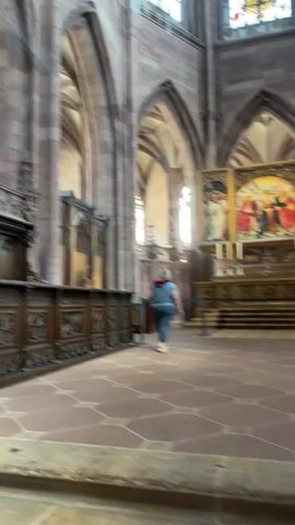This media file depicts the interior of a grand Gothic cathedral, likely Freiburg Minster, given the location context of Freiburg im Breisgau, Germany. The scene is an awe-inspiring view looking upwards towards the ornate vaulted ceiling, punctuated by large, arched stained-glass windows. Sunlight streams through these windows, creating dramatic light rays that illuminate the vast interior space.

The primary subjects are the architectural elements of the cathedral. We see massive stone pillars and arches, characteristic of Gothic architecture, supporting a complex ribbed vault ceiling. The stained-glass windows are filled with colorful patterns, suggesting they depict religious scenes or figures.

In the lower half of the frame, a person is visible walking away from the camera towards an altar area. The altar is adorned with a large triptych painting, featuring religious figures, and flanked by candles. Another person is seen in the background, also facing the altar. The floor is tiled with large, geometric stone patterns. The overall atmosphere suggests a quiet, reverent moment within a place of worship. The lighting, with the sunbeams, implies it's daytime, likely with clear skies outside to allow such strong light penetration. The presence of visitors indicates it is open to the public. There is no visible text in the media file.
