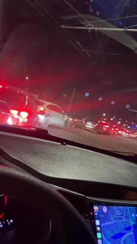 The video shows a view from inside a car at night, looking out through the windshield. The car is in traffic, with multiple cars visible ahead. The brake lights of the cars in front are illuminated red, indicating they are stopped or moving slowly. The scene appears to be on a city street, with streetlights and overhead wires visible. The dashboard of the car is partially visible, with a screen displaying what appears to be a navigation system. The lighting is dark, with the primary illumination coming from the vehicle lights and streetlights, creating a red and blue hue on the scene.