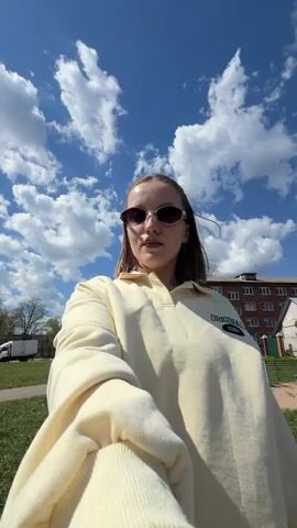 A young woman wearing sunglasses and a light yellow sweatshirt poses for a selfie video outdoors. She is standing on a paved surface in front of a grassy area with trees and buildings in the background. The sky is blue with fluffy white clouds. The woman adjusts her hair and looks at the camera. There is a playground and some trash bins visible behind her.