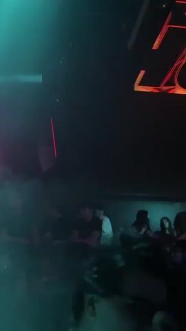The video shows a dance club with a DJ booth, a dance floor with several people and an elevated viewing platform. The DJ is wearing a white shirt. The lighting is multicolored, with a smoke machine creating a hazy atmosphere. The video includes quick panning shots across the crowd and the illuminated ceiling.