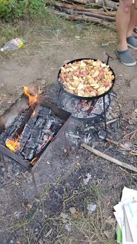 A video clip shows a close-up view of an outdoor cooking scene. In the foreground, a metal brazier contains burning logs that emit flames. To the right of the brazier, a large round pan filled with sliced potatoes is elevated on a tripod over a separate fire. The potatoes appear to be cooking, with some showing signs of browning. A person's legs are visible in the upper right corner, wearing shorts and grey slip-on shoes. They are holding a bottle of beer and a wooden utensil, suggesting they are tending to the food. The ground is covered with dirt, dry leaves, and scattered cigarette butts, indicating a rustic, possibly camping or picnic setting. In the background, a pile of cut logs lies on the ground, and foliage can be seen. A plastic bottle is on the ground to the left. The overall impression is of a casual, outdoor meal preparation.