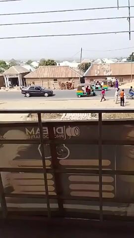 The video shows a street scene in what appears to be a small town or village. The shot is taken from an elevated perspective, looking out over a street intersection. There are several small buildings with corrugated metal roofs lining the street. A few people are walking along the side of the road, and there are also a few vehicles, including a black car and a small, three-wheeled vehicle that looks like an auto-rickshaw. The sky is overcast, and the overall impression is of a dusty and somewhat impoverished area. In the foreground, the railing of a balcony or window is visible, partially obscuring the view. A sign with the words "MAI TEAM PHOTOGRAPH" is also visible, suggesting the location may be related to a photography business.