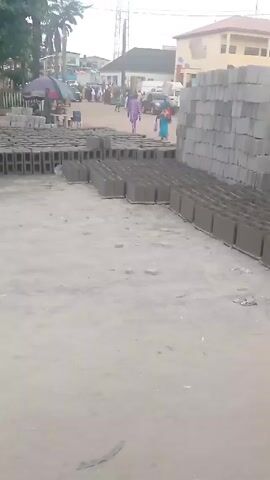 The video shows a scene at a block industry or building material sales location. Multiple stacks of gray concrete blocks sit on a sandy or dirt surface, arranged in rows. Two men wearing hats are working near the blocks, possibly moving or arranging them. People are walking in the background, past a line of shops and stands covered with tarps or umbrellas. Buildings and trees are visible in the background, indicating the location is likely a commercial or residential area.