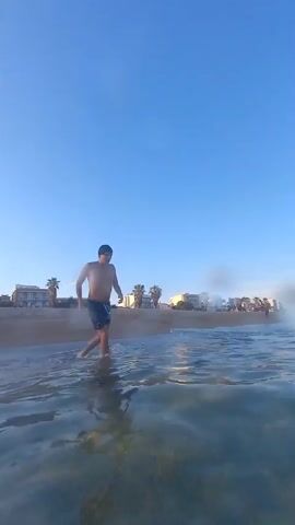 A person enters the water from a beach and swims underwater. The scene is filmed from a low angle, close to the water's surface. The water is clear and blue, and the sky is also clear and blue. In the background, there is a beach with some buildings and palm trees. A construction crane is visible on the left side of the frame. The person is wearing dark shorts and appears to be swimming with a freestyle stroke. Bubbles are visible rising from the person as they swim.